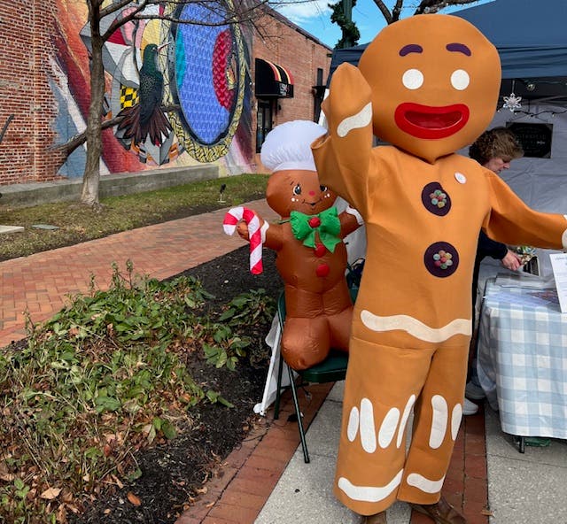 Person in gingerbread man costume, standing by a table outside and waving