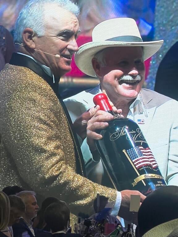 A man in a gold sparkling jacket and a man in a white suit, holding a 6 liter bottle of wine with a red white and blue American flag etched into the bottle