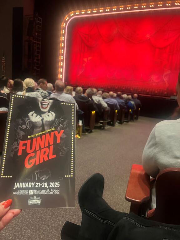 A theater full of people with a red curtain on the stage, a woman's black boots and a Funny Girl program in hand. Opening night as Funny Girl opens in Fort Myers