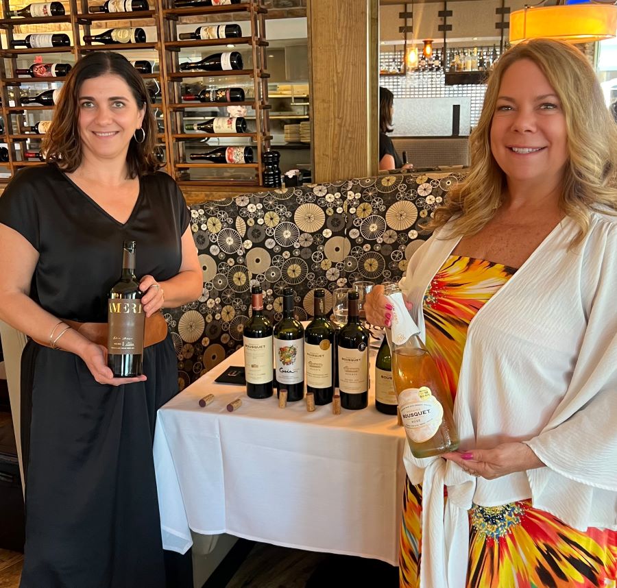 Anne Bousquet and Gina Birch posing with a bottle of wine in a restaurant getting ready for Chardonnay all day in Florida.