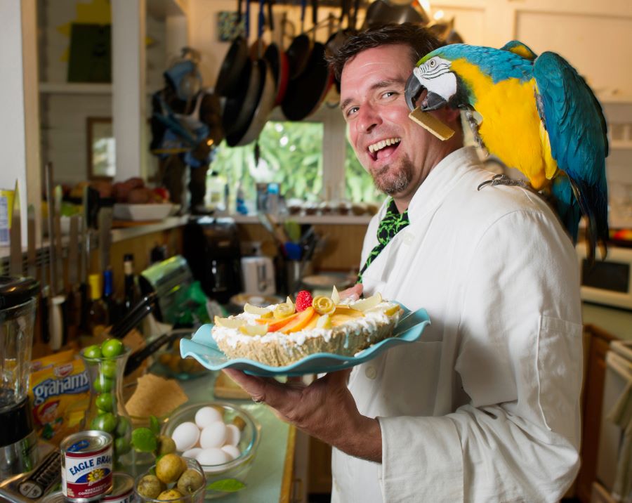 A man in white chefs coat with a blue parrot on his shoulder. The man is smiling and holding a full key lime pie at the Key West Key Lime Festival
