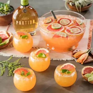 Bowl of light orange liquid with floating grapefruit slices and glasses full of the cocktail surrounding it