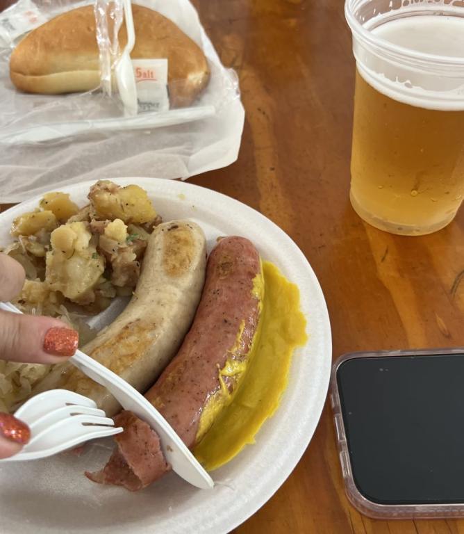 plastic plate with two sausages and a womans hand cutting one with a fork and knife, a beer in the background, from Oktoberfest leads SWFL weekend events