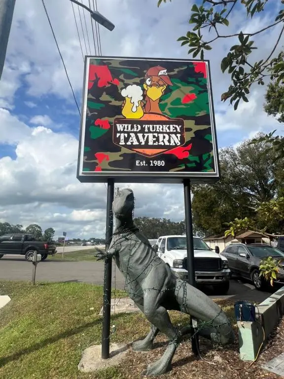 a giant statue of a dinosaur holding on to a sign by the side of the road. It reads Wild Turkey Tavern